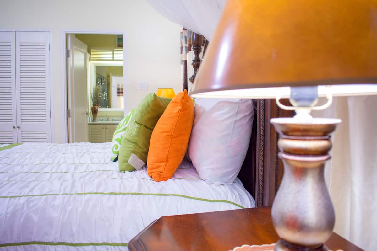 A neatly made bed with a white and green striped bedspread, a wooden nightstand with a lamp, and a mirror reflecting a bathroom with a sink and a vase.