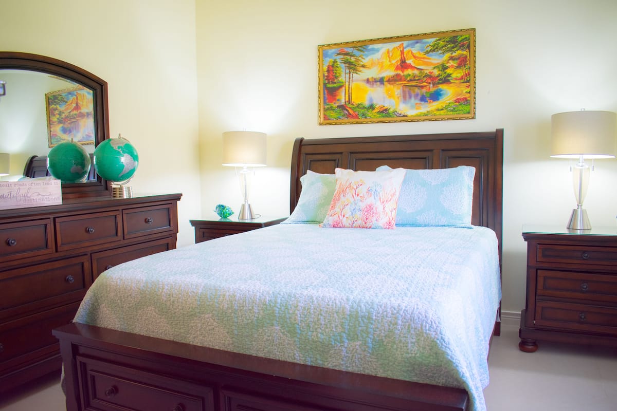 A neatly made bed with a floral bedspread, a wooden nightstand with a lamp, and a dresser with a globe and a sign.