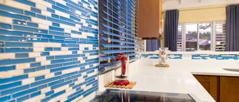 A kitchen with a blue and white tiled backsplash, a black countertop, and a window with curtains.