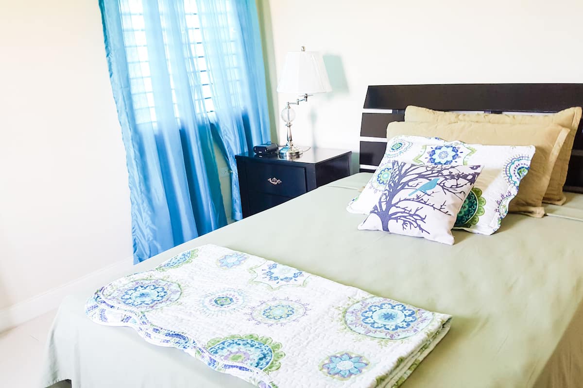 A neatly made bed with a patterned bedspread and pillows, a bedside table with a lamp, and a window with blue curtains.