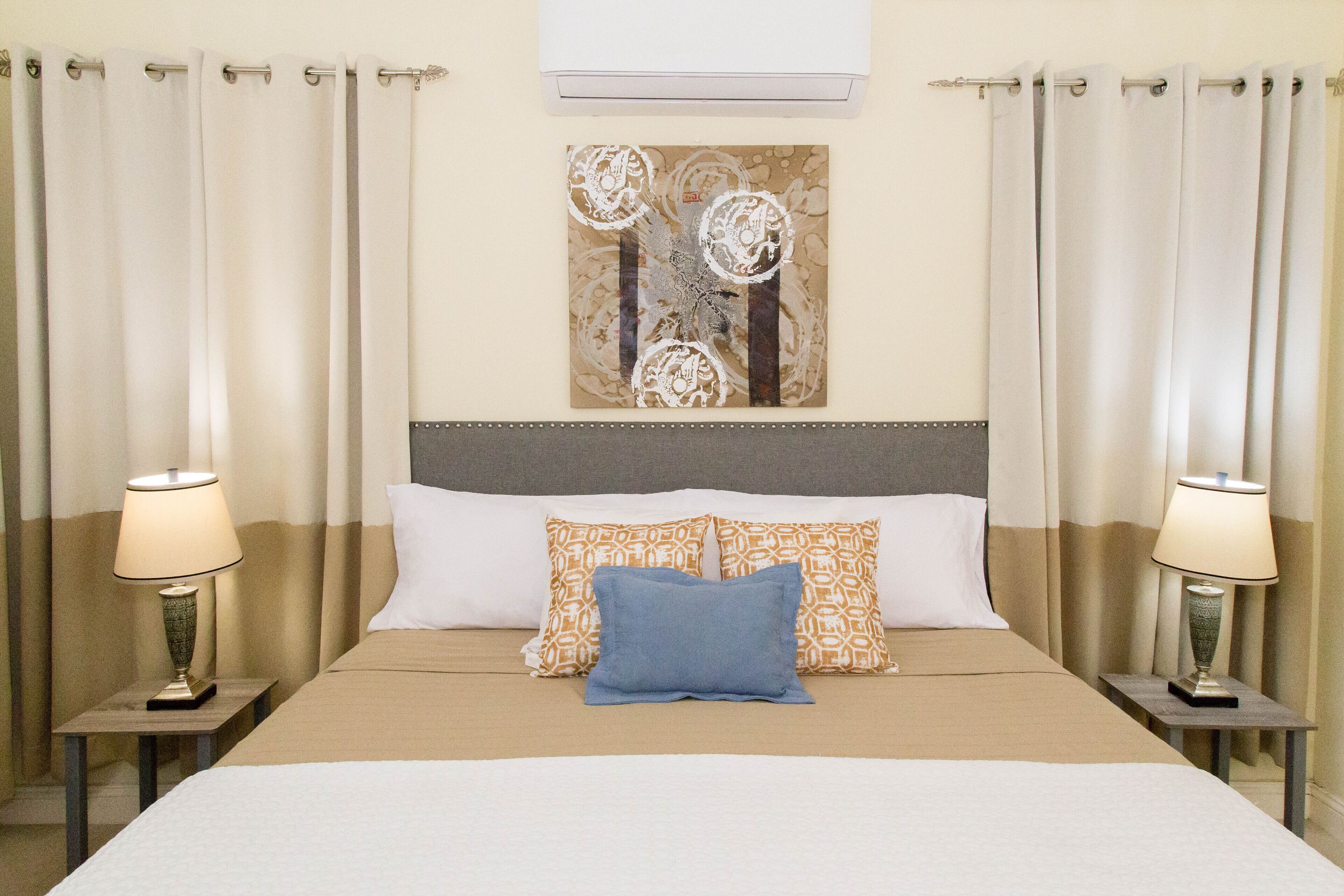 A neatly made bed with a beige bedspread, two patterned pillows, and a blue accent pillow. There are two bedside lamps on either side of the bed. A decorative wall piece hangs above the bed.