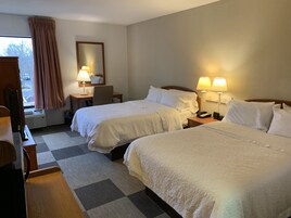 Quarto de hotel com duas camas, uma escrivaninha, uma cadeira, um abajur e uma janela com cortinas.