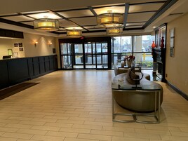 Um hall amplo com balcão de recepção, mesa de centro de vidro e vista para o estacionamento através de grandes janelas.