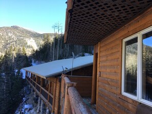 A wooden cabin with a balcony overlooking a snowy landscape and forested mountains.