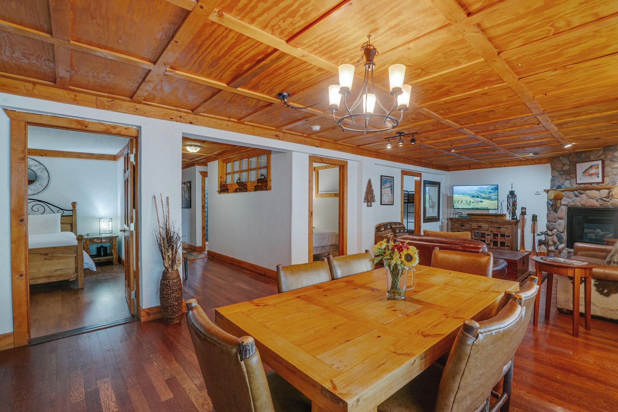 A spacious room with a wooden dining table, chairs, and a chandelier. There is a fireplace and a television mounted on the wall.