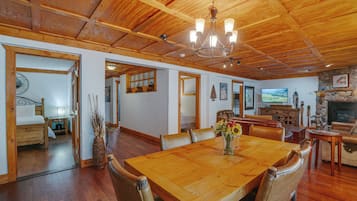A spacious room with a wooden dining table, chairs, and a chandelier. There is a fireplace and a television mounted on the wall.