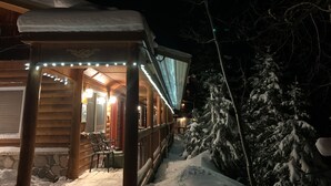 En stuga byggd av trä med en inglasad veranda, ett snötäckt tak och ett snötäckt träd i bakgrunden.