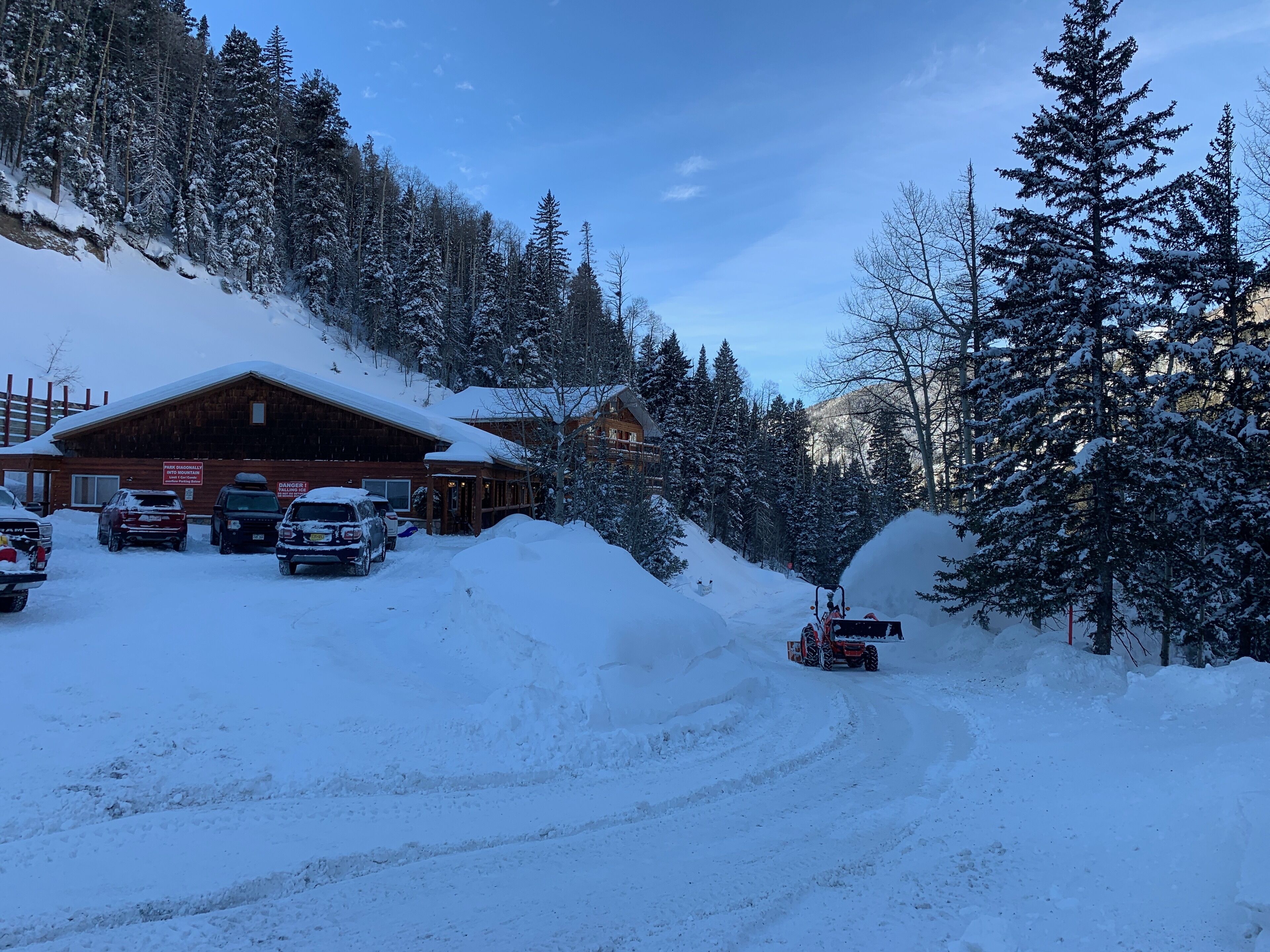 A snow-covered lodge with a wooden exterior, a parking area with several vehicles, and a snowmobile trail.