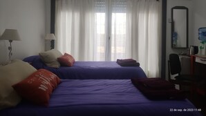 A hotel room with two beds, a desk with a chair, a large window with curtains, an air conditioning unit, and a water dispenser. - Room in homestay (Villa Real)