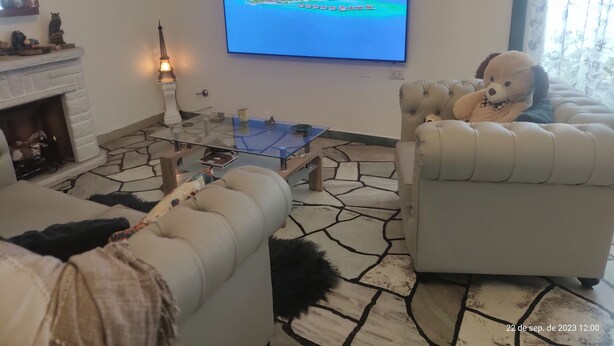 A living room with a stone-patterned floor, a glass coffee table, a fireplace, and a television displaying a tropical island. - Room in homestay (Villa Real)