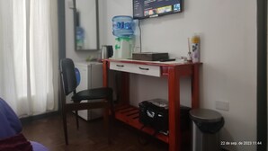 A hotel room with a desk, chair, water dispenser, and a TV mounted on the wall displaying a news channel. - Room in homestay (Villa Real)