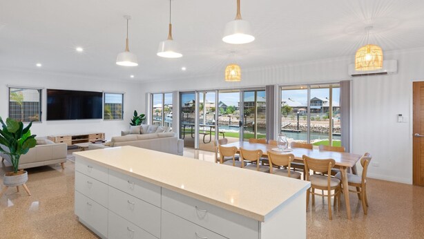 Una cocina moderna con una isla grande, comedor con sillas y vistas a una piscina y casas a través de amplios ventanales.