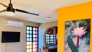 A room with a dining area, a TV, an air conditioner, and a painting of a cactus with a flower. - Casa Edilia - Smack in the middle of town/one block from the beach (Los Barriles)
