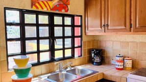 A kitchen with wooden cabinets, a sink, a window with a view, and a colorful wall decoration. - Casa Edilia - Smack in the middle of town/one block from the beach (Los Barriles)