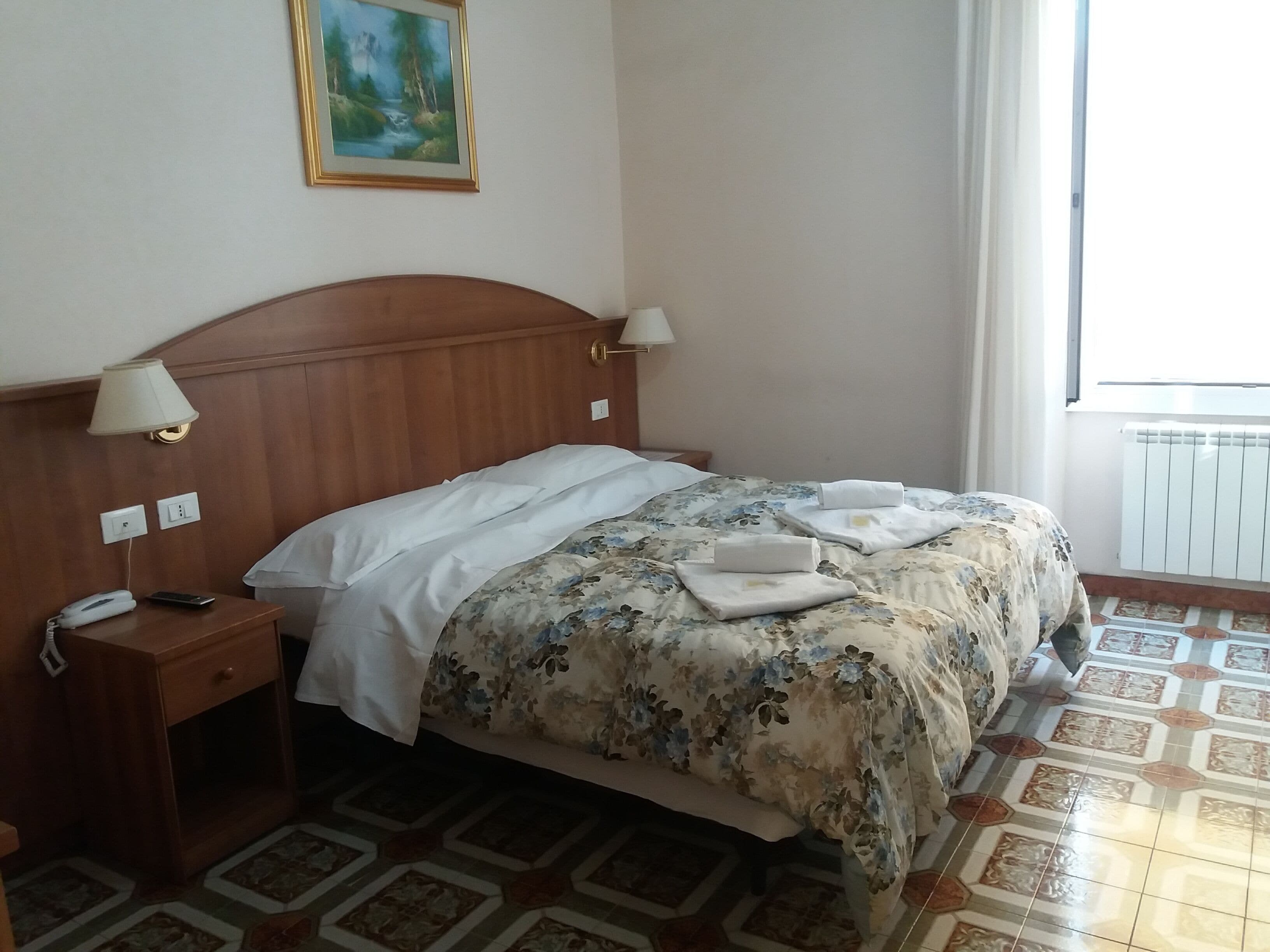 A hotel room with a wooden headboard, two bedside lamps, a telephone, a floral bedspread, and a painting on the wall.