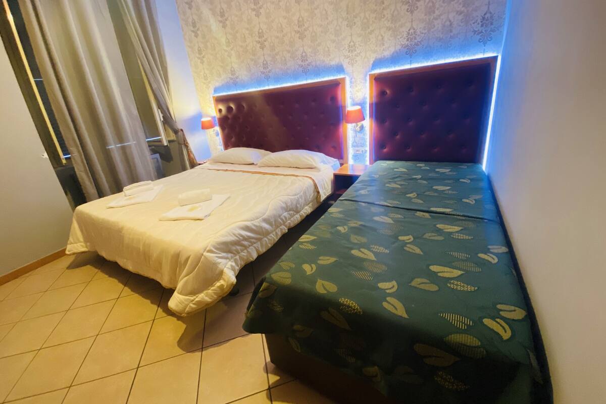 A hotel room with two beds, a patterned bedspread on one bed, a plain bedspread on the other, and a patterned wall.
