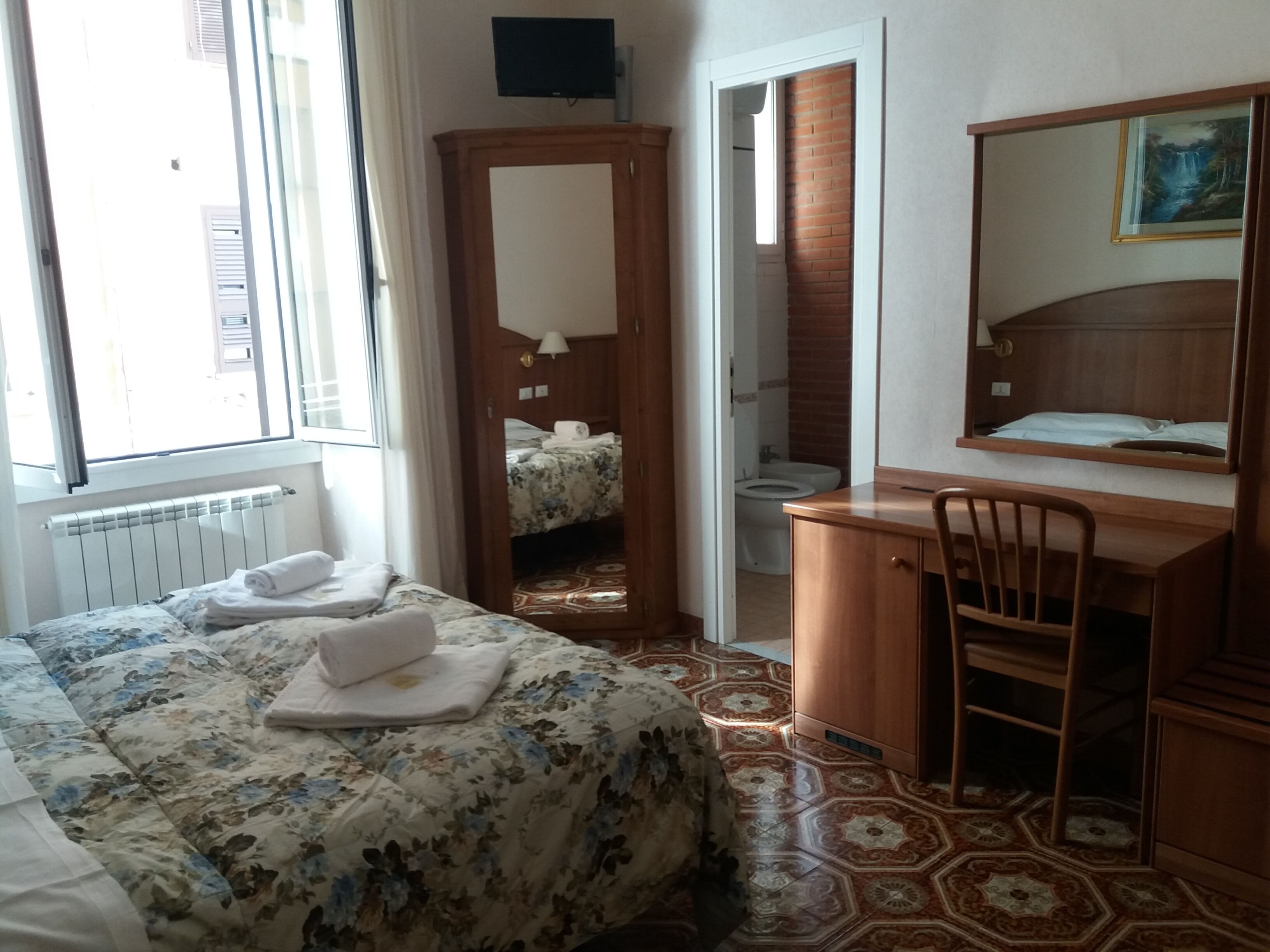 A hotel room with a bed, a desk, a chair, a bathroom, and a painting on the wall.