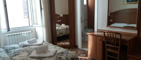 A hotel room with a bed, a desk, a chair, a bathroom, and a painting on the wall.