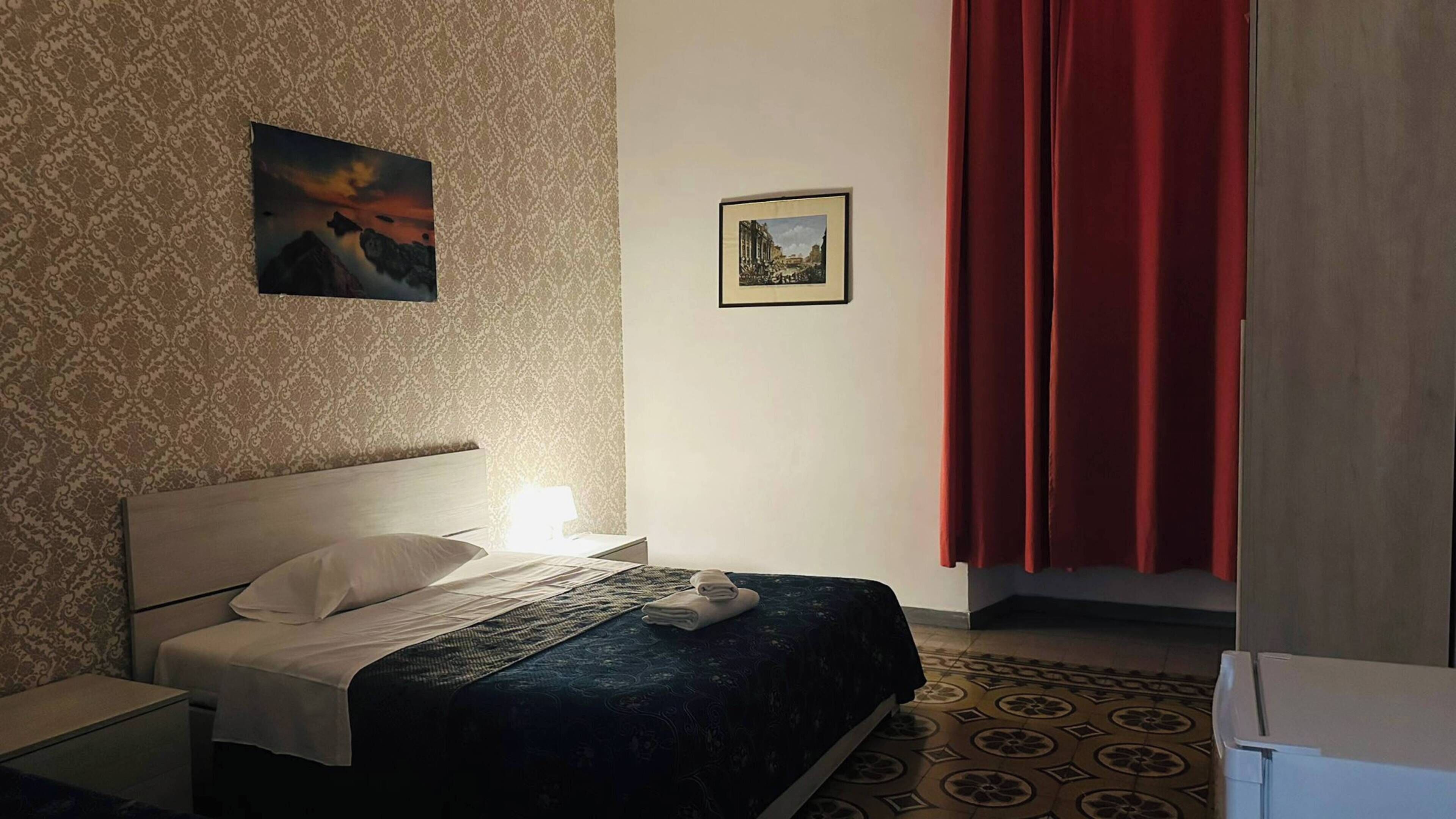 Quarto de hotel com uma cama, mesa de cabeceira, abajur, cortina vermelha e duas imagens emolduradas na parede.