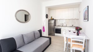 A modern living room with a grey sofa, a dining table with white chairs, a kitchen area with white cabinets, and a round mirror on the wall. - Apartments L&M - One Bedroom Apartment with Terrace (Dubrovnik)