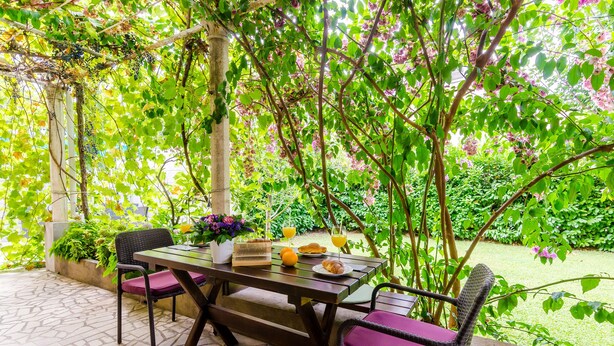 An outdoor patio with a table set for two, surrounded by lush greenery and a pergola. - Apartments L&M - One Bedroom Apartment with Terrace (Dubrovnik)