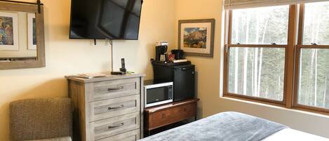 A bedroom with a bed, a TV mounted on the wall, a dresser, a chair, and a window with a view of trees.