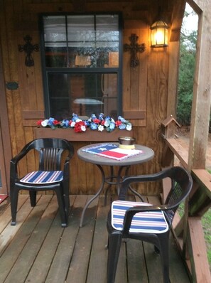 A wooden deck with two black chairs and a small round table, a window with a cross decoration, and a lantern on the wall. - Private Cabin with hot tub. Nestled in the Woods just off Lake Limestone, Texas. (Jewett)