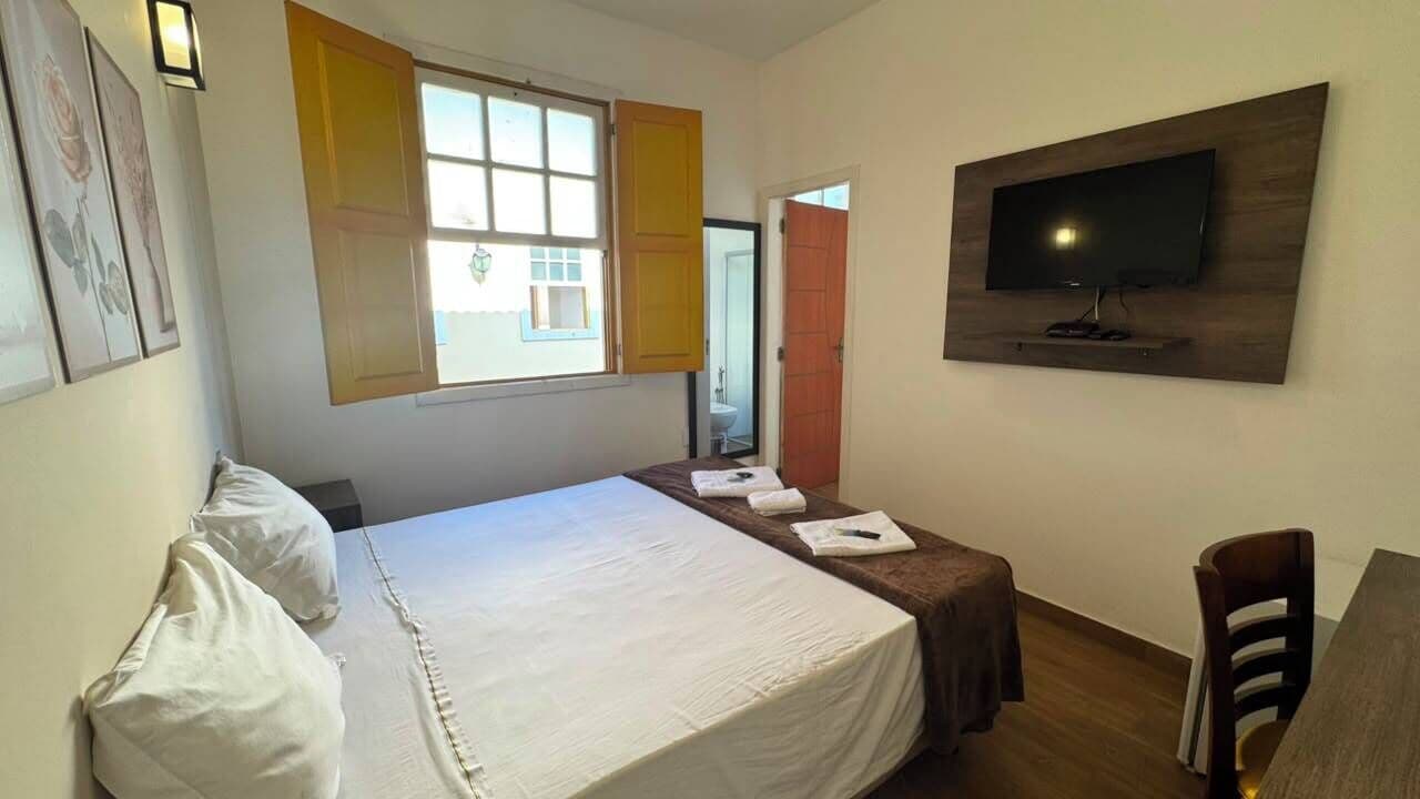 A hotel room with a bed, a TV mounted on the wall, a window with shutters, and a desk with a chair.