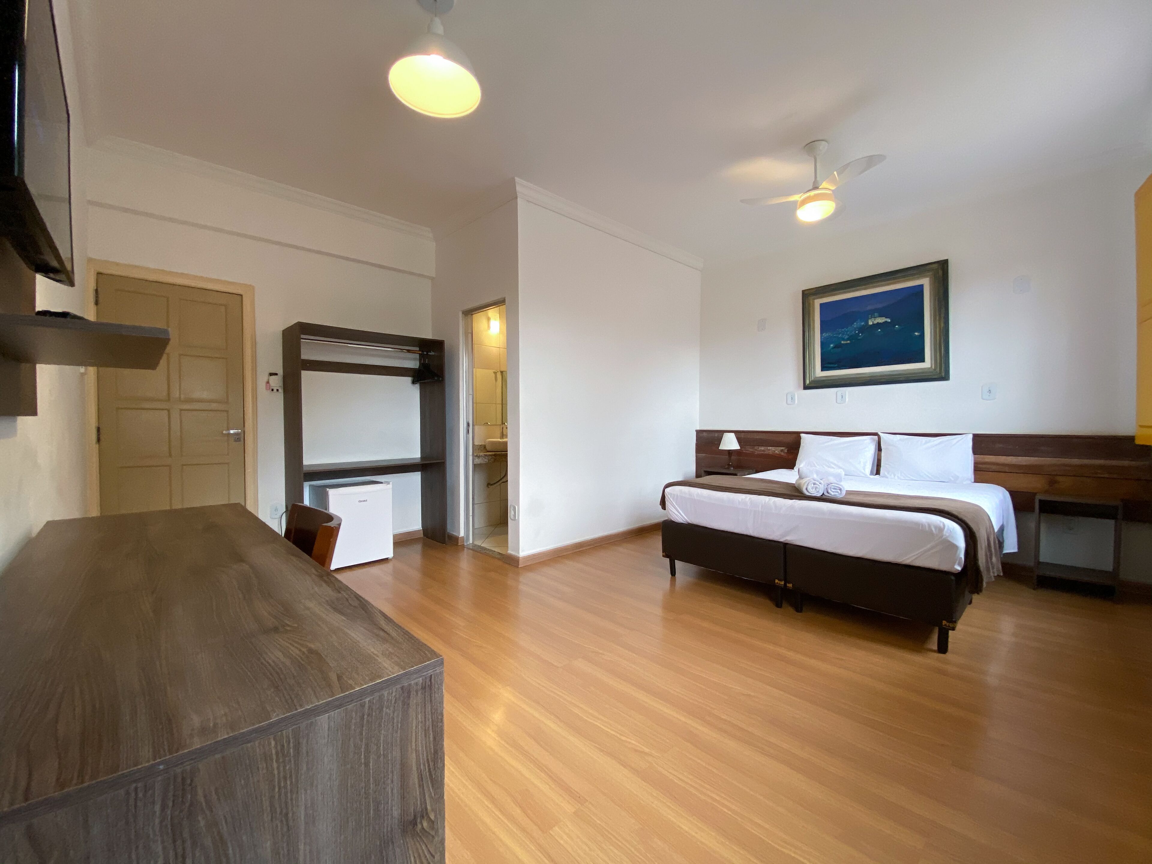 Habitación de hotel con cama, escritorio, refrigerador, ventilador de techo y un cuadro en la pared.
