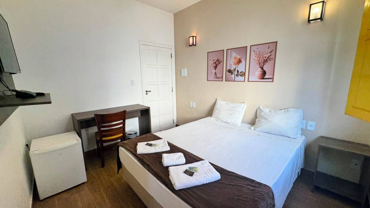 A hotel room with a bed, a desk, a chair, a small table, and a wall-mounted TV.