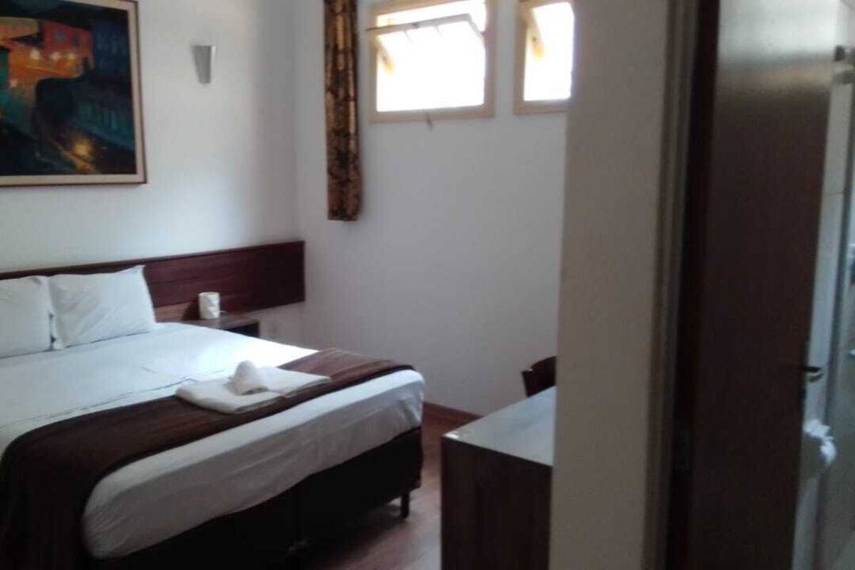 Ein Hotelzimmer mit Bett, Nachttisch, Schreibtisch, einem Bild an der Wand und einem Fenster mit Vorhängen.