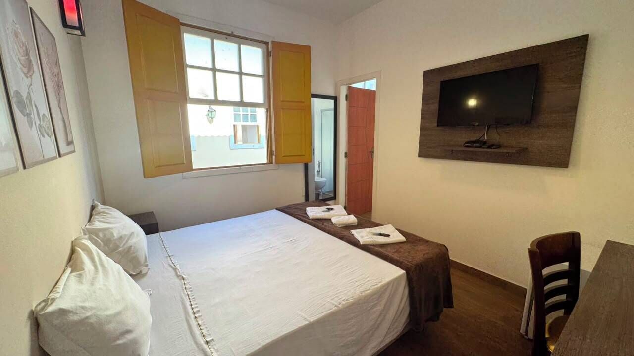 A hotel room with a bed, a TV mounted on the wall, a window with shutters, and a door leading to another room.