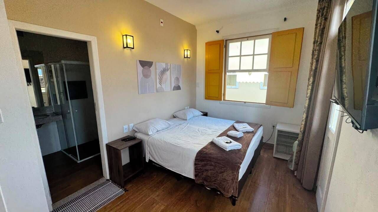Quarto de hotel com cama, mesas de cabeceira, janela com cortinas e porta de acesso a um banheiro.