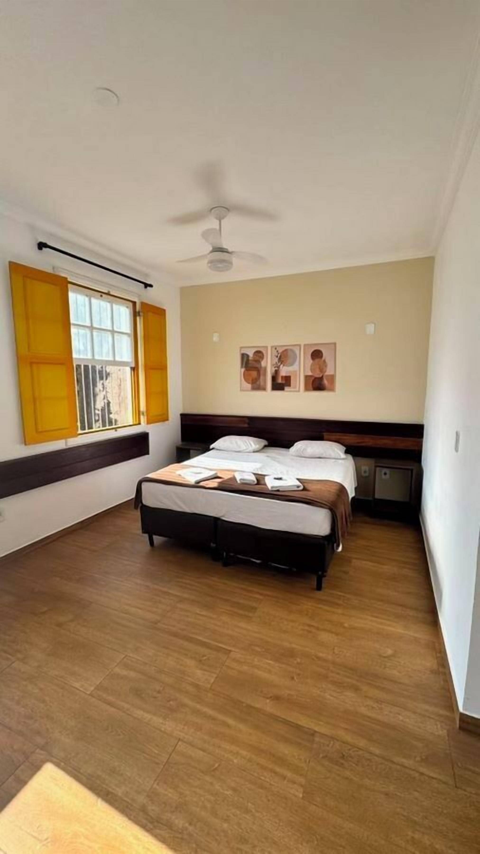 A bedroom with a bed, a ceiling fan, a window with yellow shutters, and artwork on the wall.