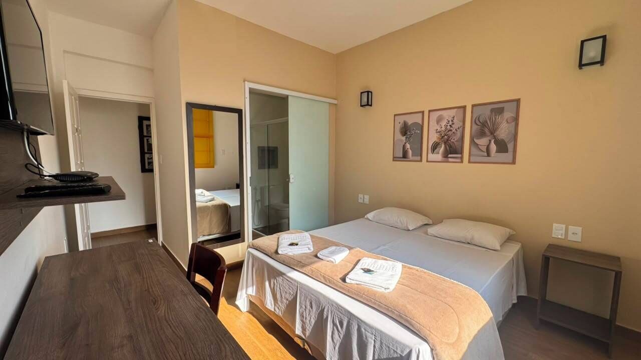 A hotel room with a bed, a desk, a chair, a wardrobe, and a bathroom visible through a glass door.