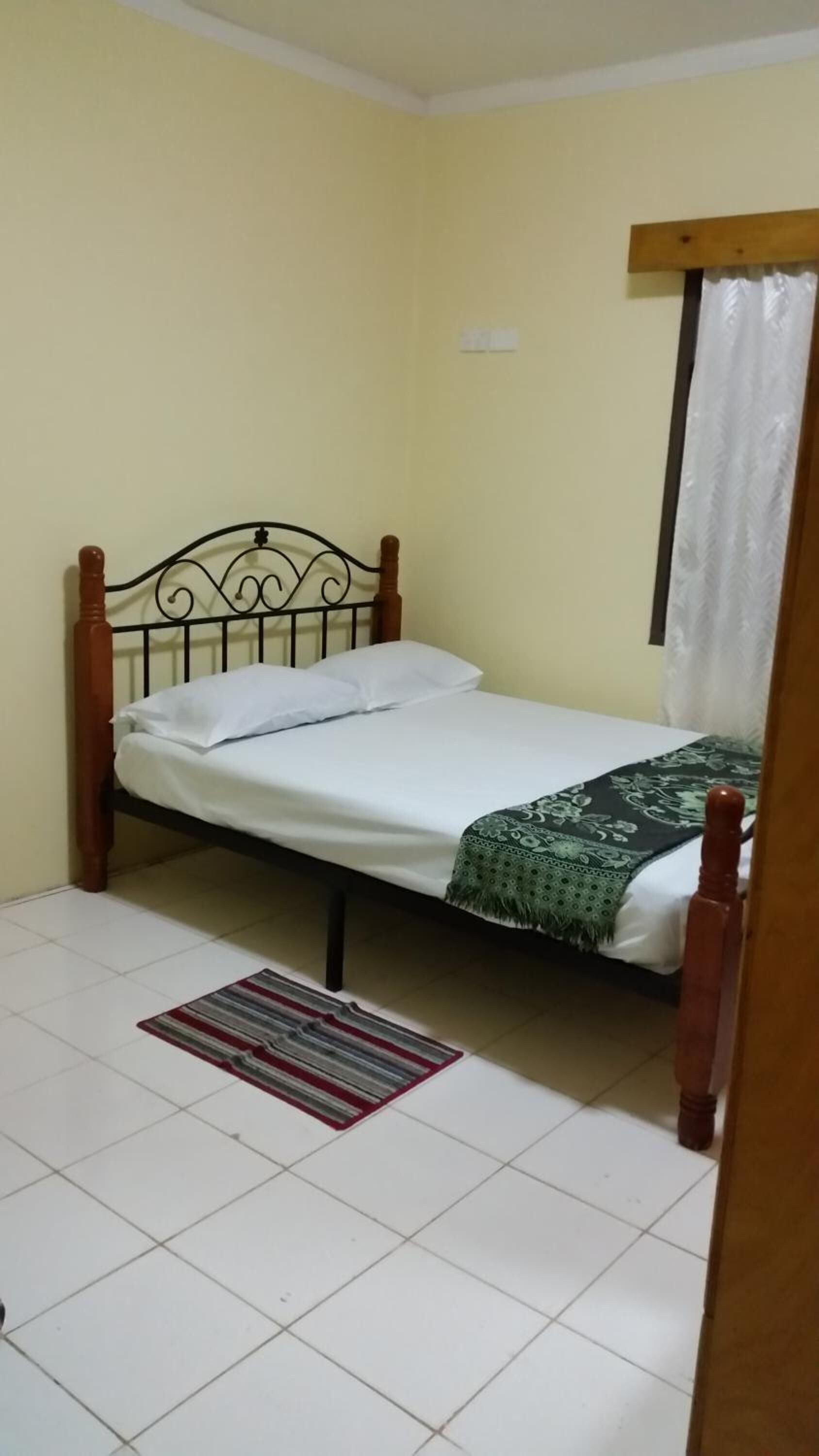 A single bed with a wooden frame and a white mattress, a green patterned bedspread, and a striped rug on a tiled floor.