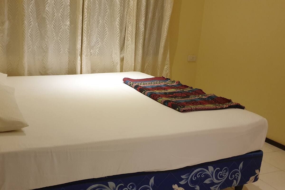 A bed with a white mattress and a colorful blanket, a white pillow, and a blue bed skirt.