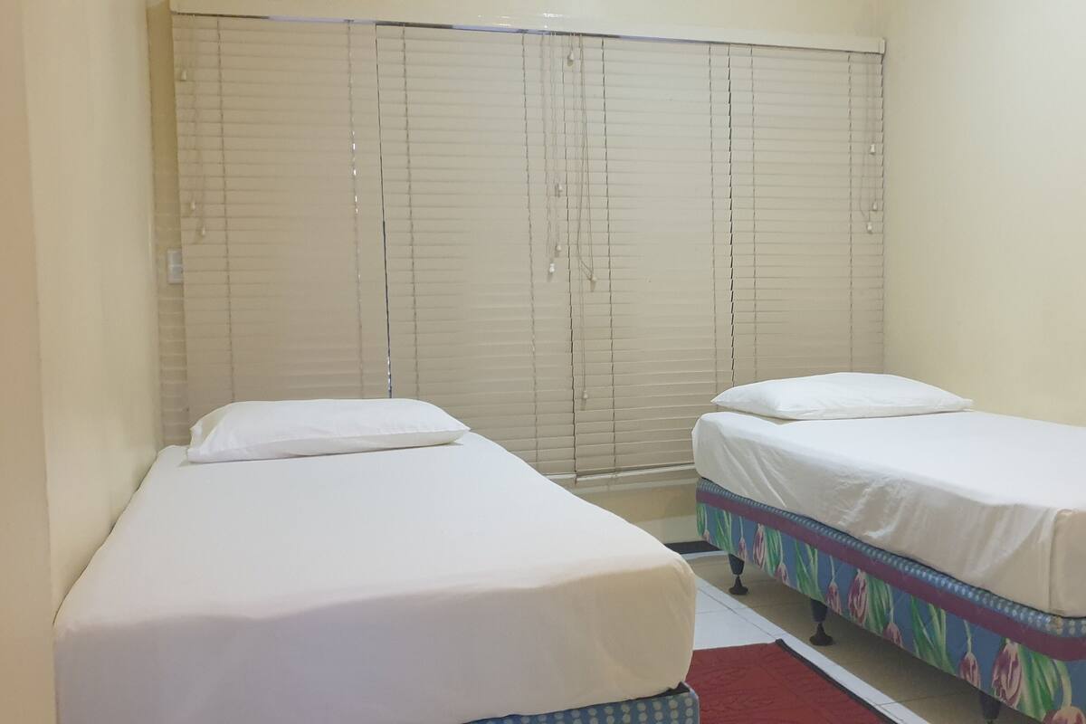 A small room with two single beds, a window with blinds, and a red rug on the floor.
