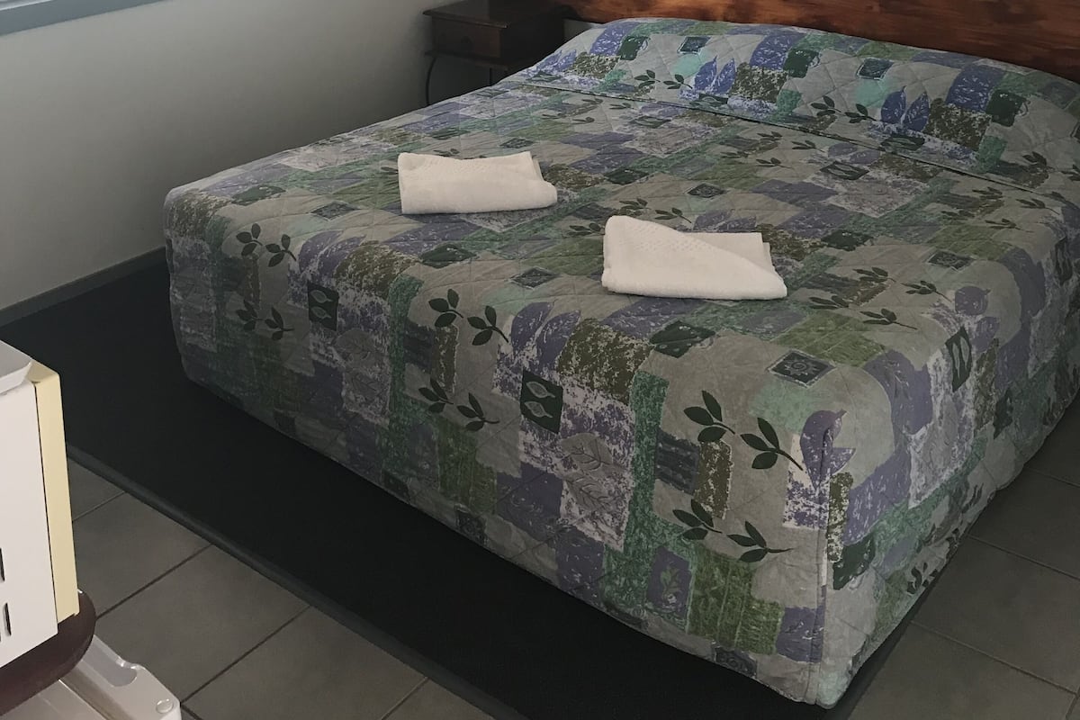 A neatly made bed with a floral-patterned bedspread and two folded towels, a window with blinds, and an air conditioning unit mounted on the wall.