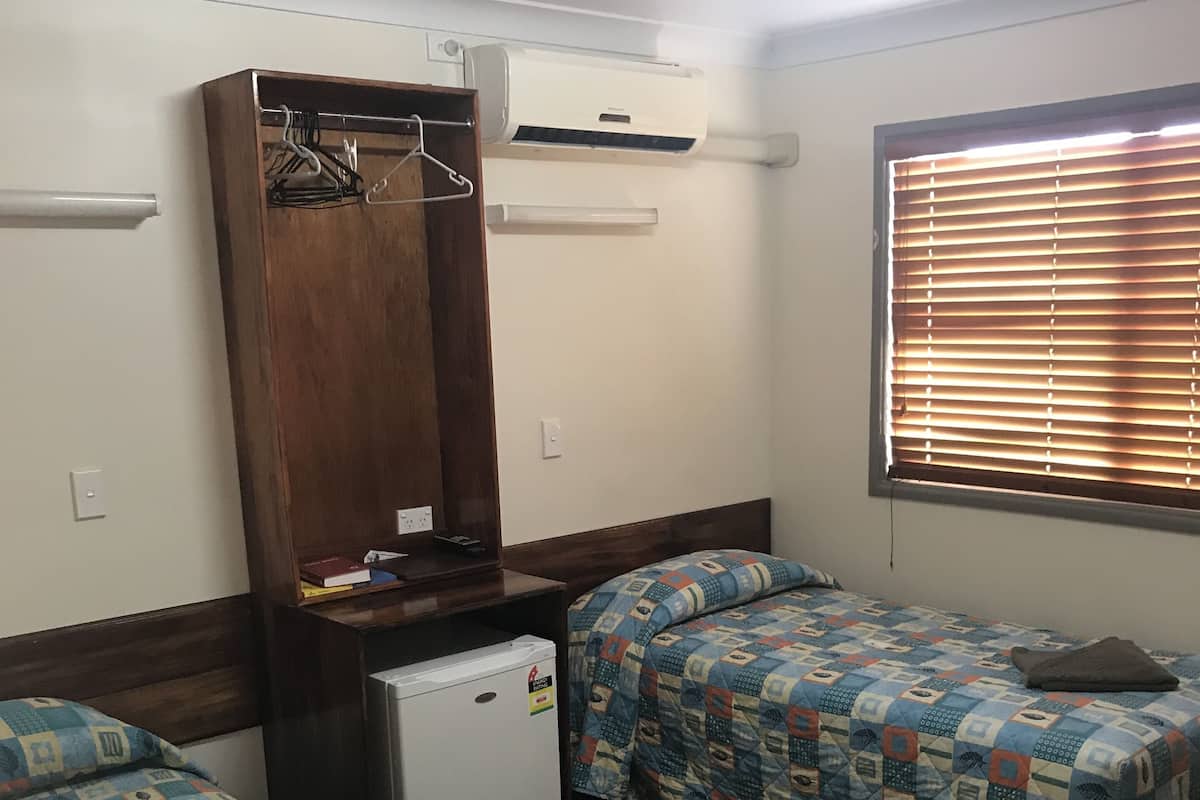 A small, compact hotel room with two beds, a small fridge, a wardrobe, and a window with blinds.