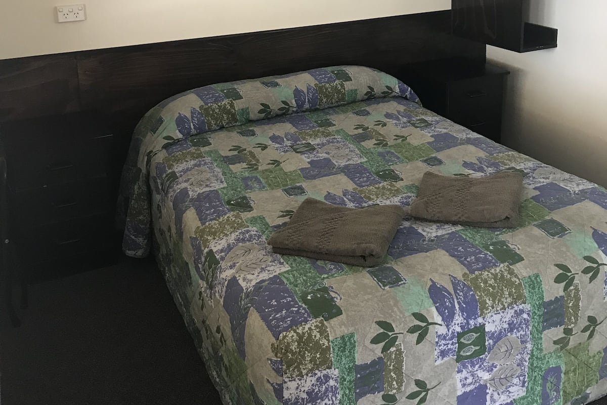 A bedroom with a bed, a dark headboard, a framed picture on the wall, and a small shelf.