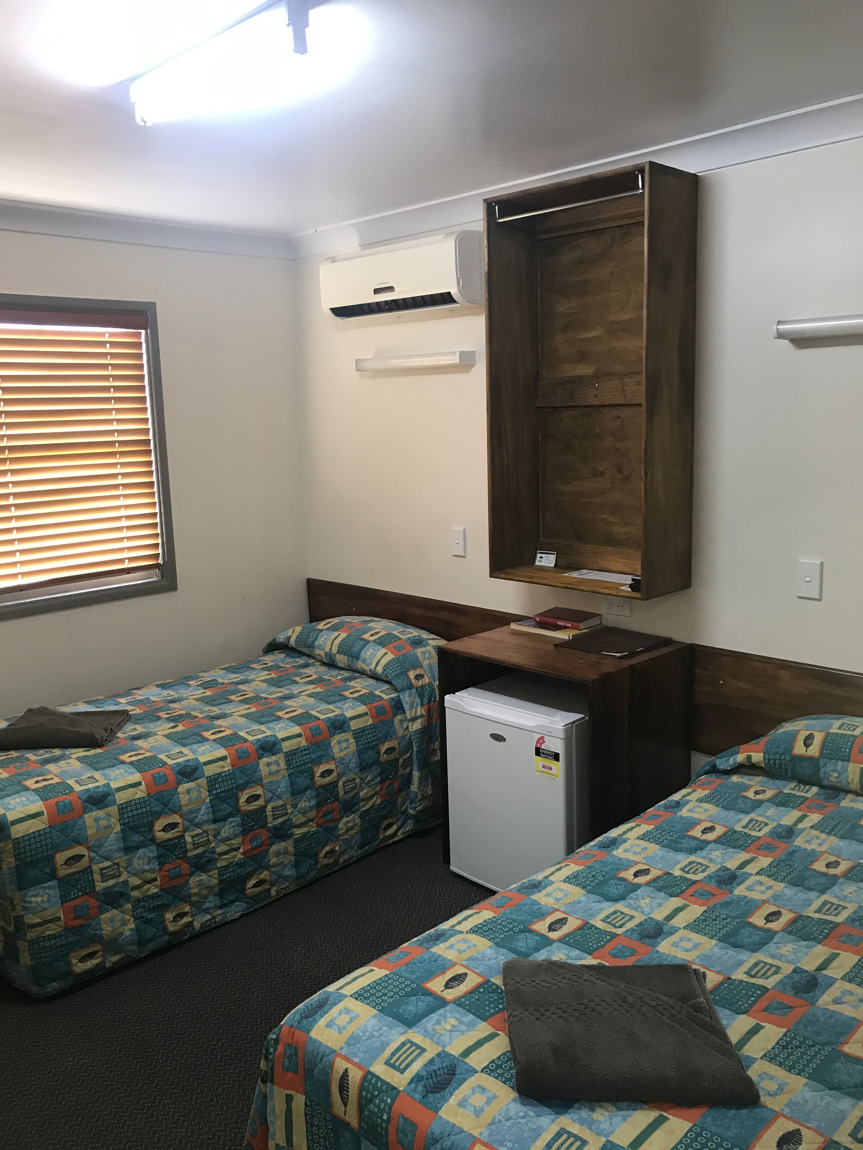 A room with two beds, a small refrigerator, a wooden wardrobe, and a window with blinds.