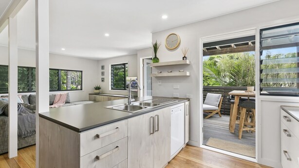 Una cocina moderna con una isla grande, un comedor con mesa y sillas de madera, y un cerramiento de vidrio corredero que da a un patio exterior con plantas.