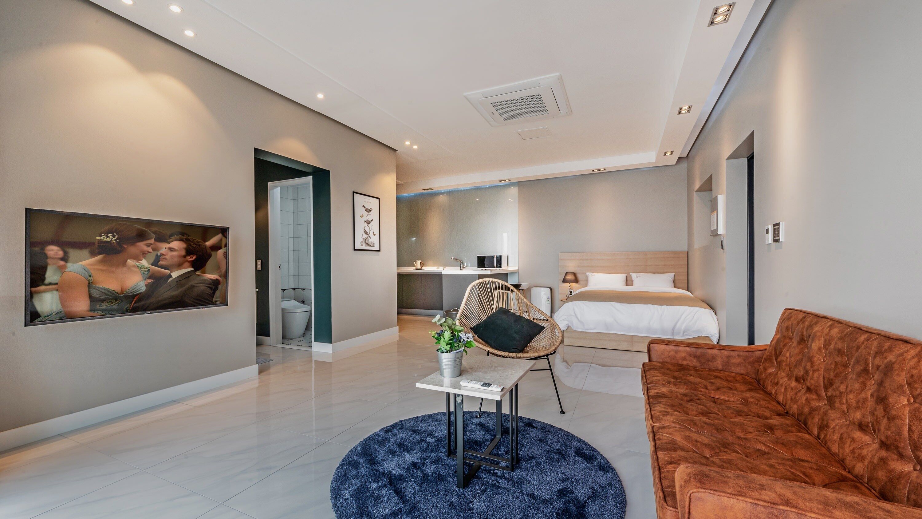 A modern hotel room with a bed, a sofa, a small table with a plant, a TV, and a bathroom with a shower and toilet.