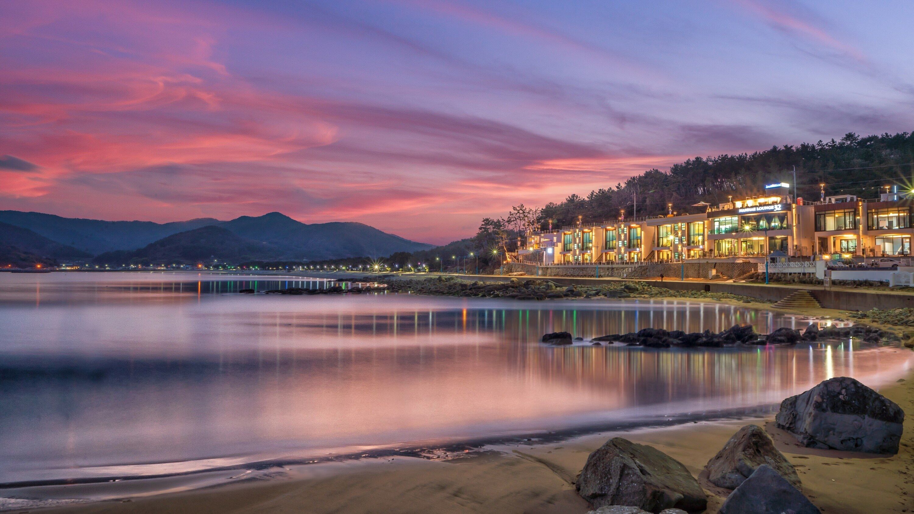 A waterfront hotel with a modern design, featuring a long reflective pool, a bridge, and a mountainous backdrop.
