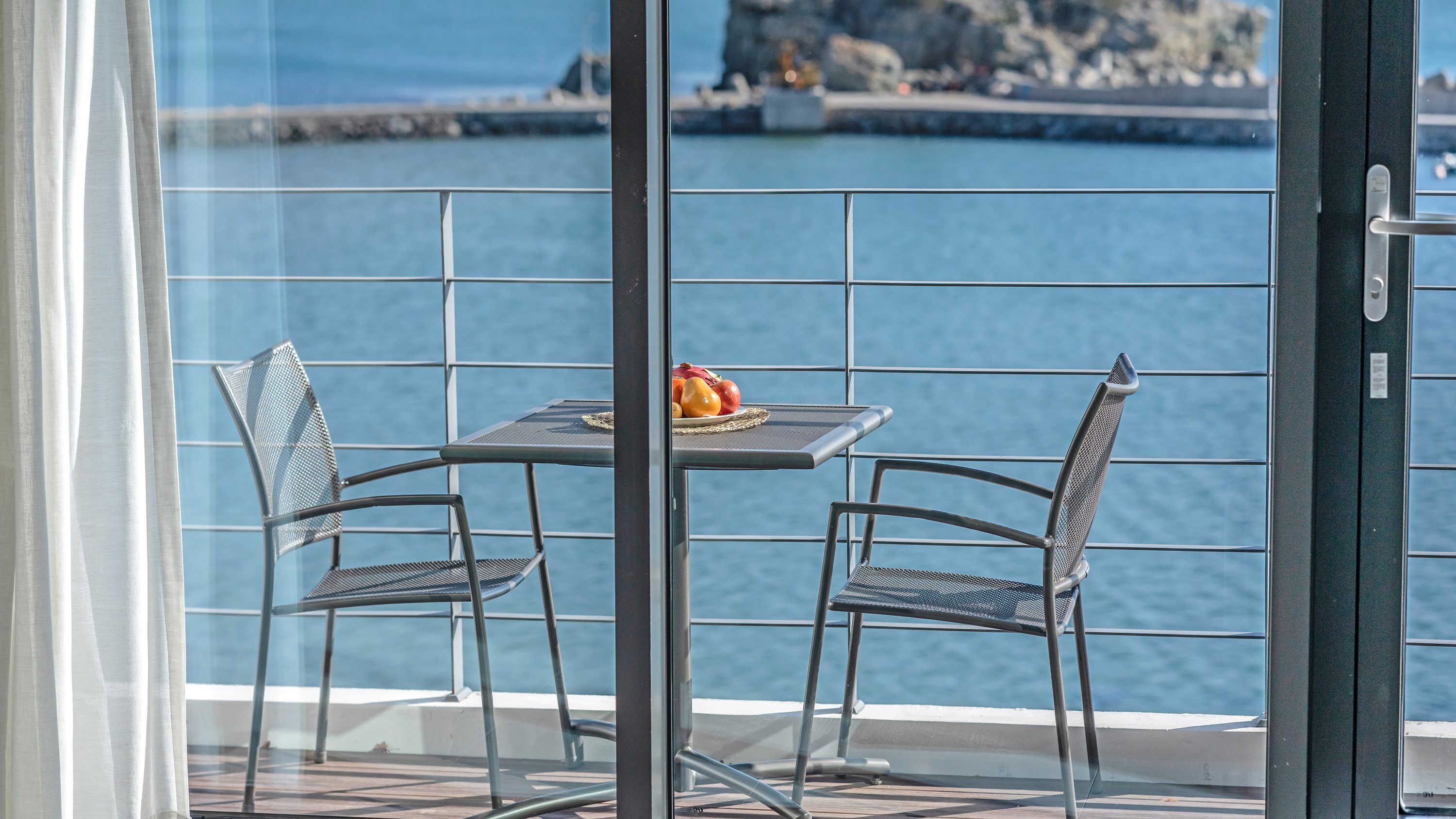 A balcony with a table and two chairs, overlooking a body of water with a rocky shoreline.