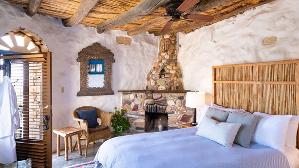 Un dormitorio rústico con chimenea de piedra, ventilador de techo de madera y una cama con colcha estampada.