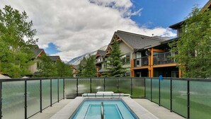A rectangular pool with a stone border, surrounded by a glass fence, set against a backdrop of houses and mountains. - Kananaskis Getaway, 1Br. Condo in the mountains (Dead Man's Flats)