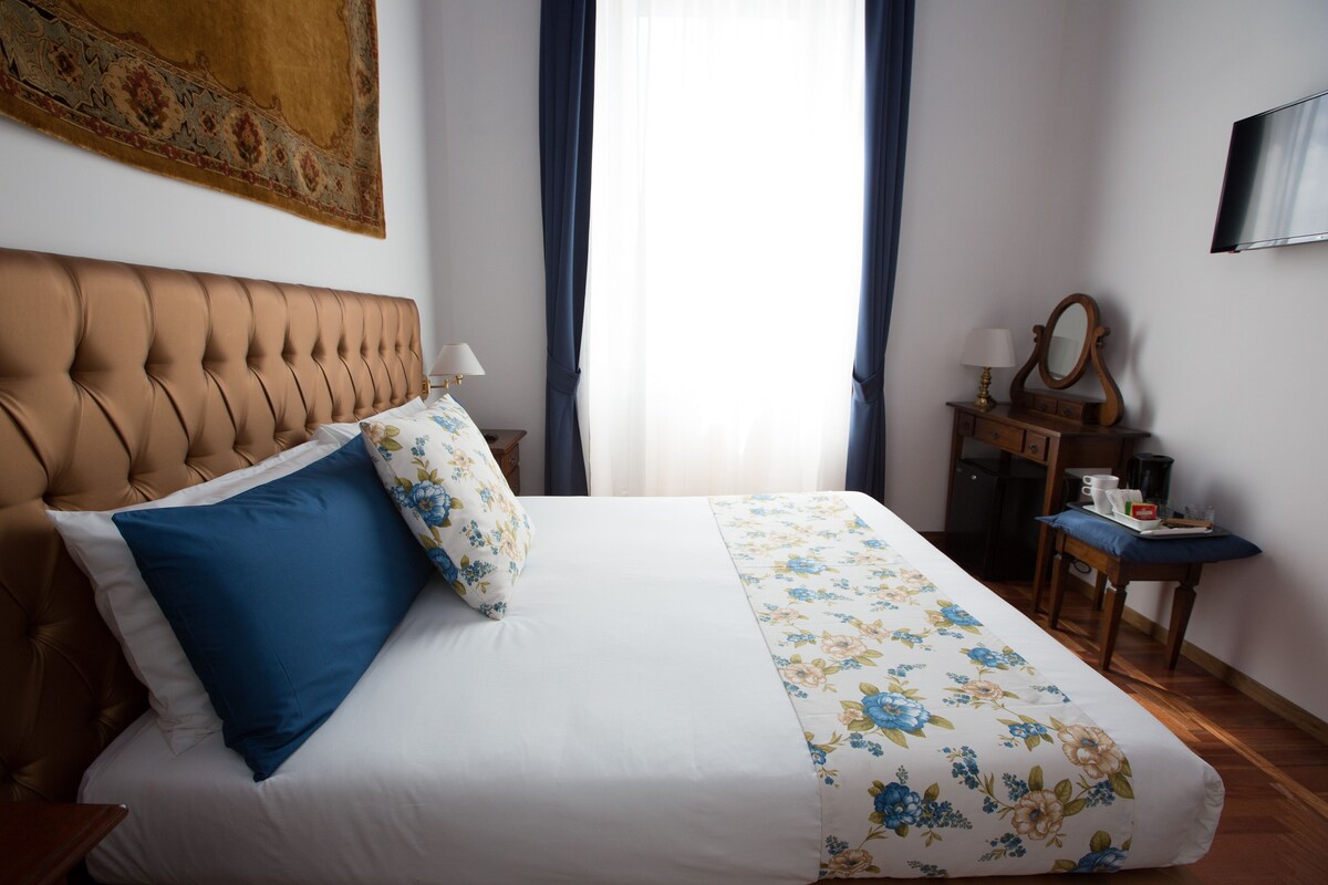 A bedroom with a tufted headboard, a bed with floral bedding, a bedside table with a lamp, a mirror, and a flat-screen TV on the wall.