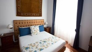 A bedroom with a tufted headboard, a floral bedspread, a nightstand with a lamp, and a large window with curtains. - Ad 2015 Guesthouse (Rome)